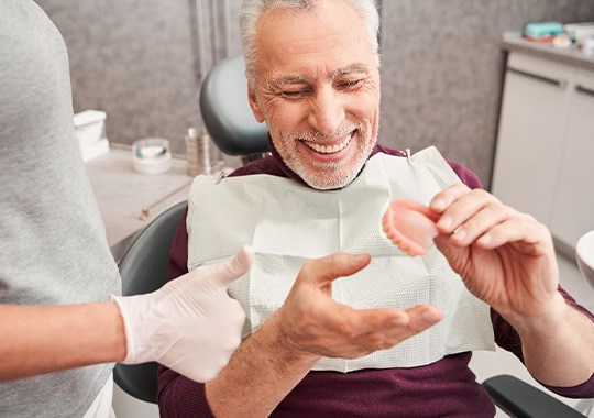 Man in dental chair smiling holding arch of dentures